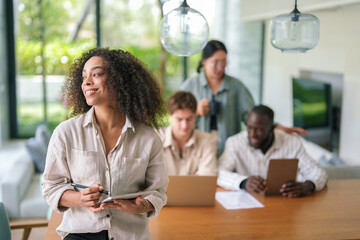A young African American businesswoman appears content while working on her digital tablet, surrounded by diverse colleagues in a bright, modern office setting.