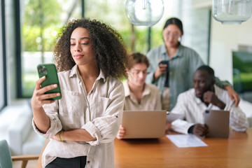 A cheerful African American businesswoman engages with her smartphone, surrounded by diverse colleagues working on laptops in a well-lit modern office setting.