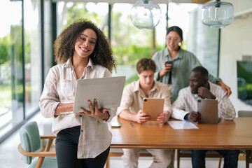 A joyful African American businesswoman works on her laptop, standing confidently in a modern office, with diverse colleagues engaged in tasks behind her.