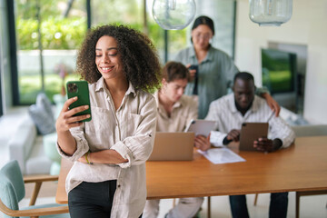 A cheerful African American young adult businesswoman is engaging with her smartphone, possibly messaging, amidst diverse colleagues focused on their laptops in a well-lit, stylish office space.
