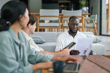 A group of young adults of various ethnic backgrounds engage in discussion while looking at documents, surrounded by laptops and smartphones in a casual office setting.