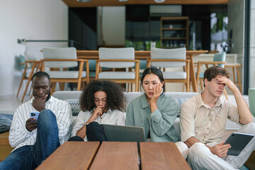 A thoughtful group of young diverse executives engage with digital devices in a casual office setting, reflecting modern collaborative work culture.
