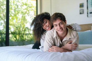 A cheerful African American woman and a smiling Caucasian man are lying on a bed, sharing a joyful moment in a bright, cozy room.