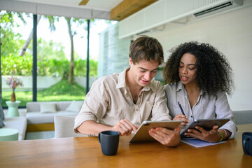 African American woman and Caucasian man in casual attire engage with a digital tablet while sitting at a wooden table, with a bright home interior as the backdrop.