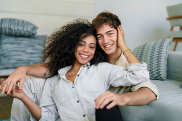 An African American woman and a Caucasian man share a relaxed, joyful interaction while sitting closely on a sofa, dressed casually in comfortable attire, in a well-lit living space.
