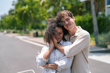 A joyful African American woman and a delighted Caucasian man in casual attire enjoy a close hug on a sunny suburban street.