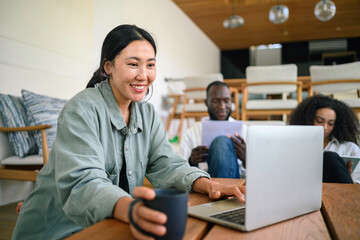 A joyful Asian female young adult uses a laptop in a well-lit home office, her attire casual. A diverse group of colleagues or friends accompany her in the background.