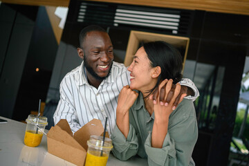 A cheerful Black man and an Asian woman enjoy a casual dine-in with drinks, sharing laughter and close company in a cozy home setting.