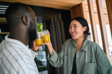 A Black man and Asian woman share smiles and a toast with healthy drinks in a cozy home setting,...