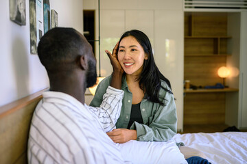 Smiling Asian woman touches the face of a Black man as they relax together in a cozy bedroom, showing casual attire and a bond of intimacy.