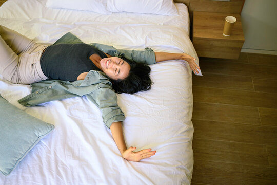 A joyful Asian woman lies spread-eagle on a bed, wearing casual attire with a content expression, surrounded by the tranquility of her well-kept bedroom.