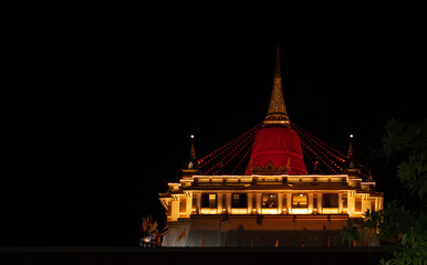 Wat Saket Ratchaworamahawihan. Temple of the Golden Mount Bangkok. A large red building with a red roof is lit up at night. The building is surrounded by trees and he is a temple.