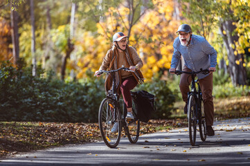 Obraz premium Joyful senior couple cycling through a scenic park on a sunny autumn day, laughing and enjoying their active outdoor lifestyle surrounded by vibrant fall colors.