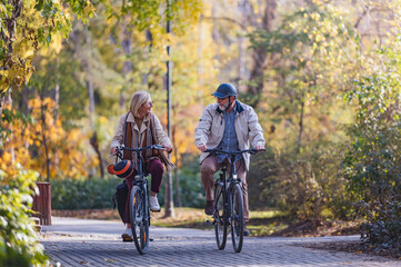Fototapeta premium Joyful senior couple cycling through a scenic park on a sunny autumn day, laughing and enjoying their active outdoor lifestyle surrounded by vibrant fall colors.