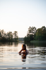 Fototapeta premium A woman swims in a calm lake at sunset, surrounded by reflections of trees and the soft colors of twilight. This peaceful scene highlights relaxation and connection with nature.