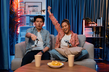 Sitting sofa of happy couple playing video game with joysticks, smiling woman playing challenge, raising fist up to be winner the challenge at modern neon blub light living room at home. Infobahn.