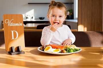 Kid girl tasting dish of salmon, eggs, mushrooms and greens. Wooden board with Vitamin D. Sunlit kitchen glow. Healthy food Concept. 