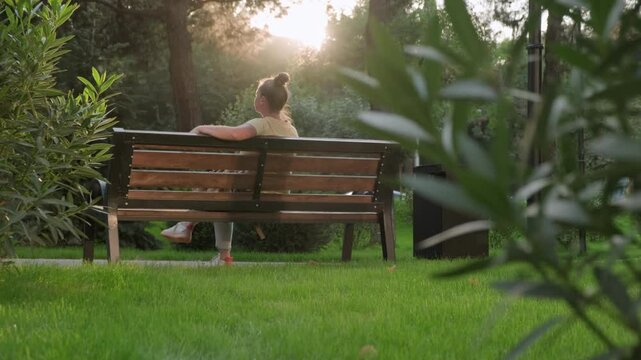 Woman relaxing sitting on bench in city park in rays of sun at sunrise in morning.