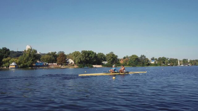Rowing team training. Side view of 2 young caucasian male rowers, during a rowing practice, athlete sitting in a boat in the river Dnipro, rows through a calm water sunny day, autumn. 4k footage