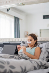 woman lies in bed in casual clothes and with bandages on her face after plastic surgery on her nose, files nails and watches favorite movie on tablet,