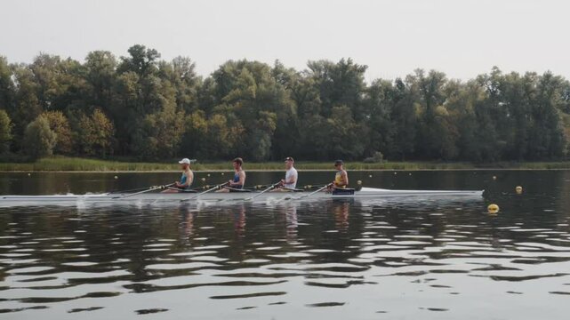 Rowing team training. Side view of 4 young caucasian male rowers, during a rowing practice, athlete sitting in a boat in the river Dnipro, rows through a calm water in autumn. 4k footage. City area in