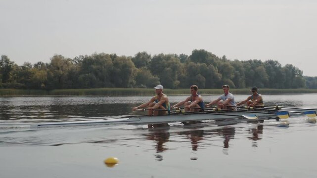 Rowing team training. Side view of 4 young caucasian male rowers, during a rowing practice, athlete sitting in a boat in the river Dnipro, rows through a calm water in autumn. 4k footage. City area in