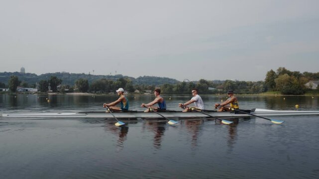Rowing team training. Side view of 4 young caucasian male rowers, during a rowing practice, athlete sitting in a boat in the river Dnipro, rows through a calm water in autumn. 4k footage. City area in