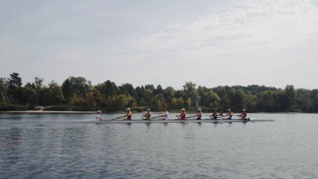 Rowing team training. Side view of 8 young caucasian male rowers, during a rowing practice, athlete sitting in a boat in the river Dnipro, rows through a calm water in autumn. 4k footage. City area in
