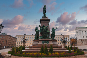 The statue of Alexander II stands in Senate Square, Helsinki, with the Helsinki Cathedral partially visible. A vibrant sky with clouds forms the backdrop.