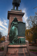 A bronze statue of a woman in classical attire leans on a stone pedestal in Helsinki, Finland. The base features books, set against a park with trees and clear sky.