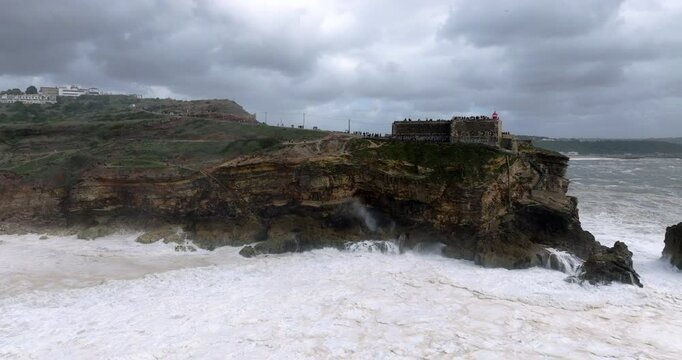 Aerial drone shot of big waves breaking on rocks and cliffs on a day with giant waves in Nazare, Portugal, Europe. Farol da Nazar&eacute; lighthouse visible. Shot in ProRes 422 HQ Log