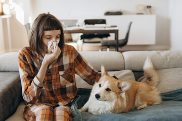 Young Woman Blowing Her Nose While Sitting On The Sofa With Her Dog, Suffering From Illness At Home