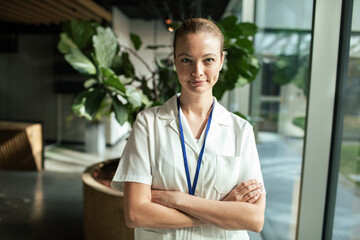 Confident female healthcare worker in white coat with arms crossed, standing in hospital lobby