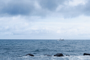Small Fishing Boat Sailing Far Away On Blue Sea With Rocks And Cloudy Sky