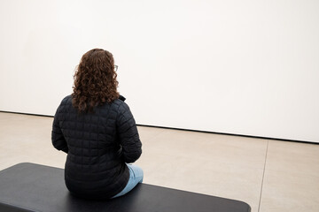 Young Caucasian Woman Sitting on a Bench in a Minimalist White Room, Contemplating