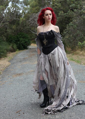 Fototapeta premium full length portrait of beautiful red haired female model figure, wearing gothic fantasy costume with torn ballgown wedding dress, black corset, leather boots. walking pose forest scenery background.