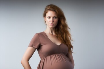 Fototapeta premium A pregnant woman standing with her back slightly arched, placing her hands on her lower back while looking towards the camera with a worried expression. She is wearing a comfortable maternity dress