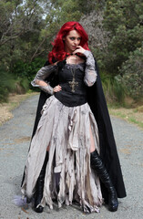 Fototapeta premium full length portrait of beautiful red haired female model figure, wearing gothic fantasy costume with torn ballgown wedding dress, black corset, leather boots. walking pose forest scenery background.