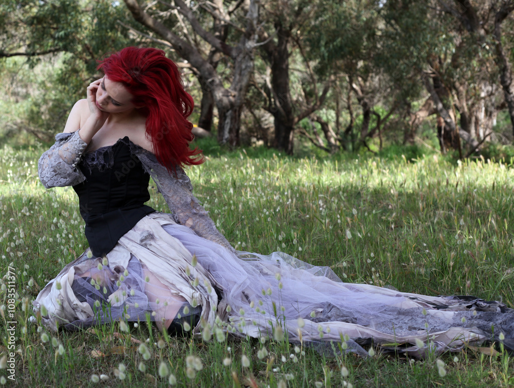 Wall mural full length portrait of beautiful red haired female model figure, wearing gothic fantasy costume with torn ballgown wedding dress, black corset, leather boots. sitting pose forest scenery background.