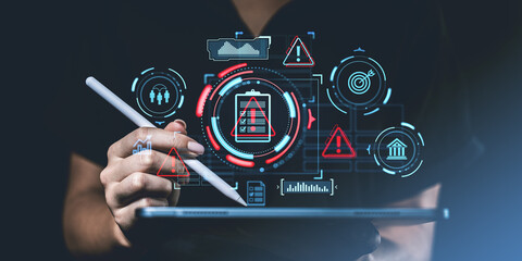 Hand holding a stylus over a tablet with futuristic risk management icons such as warning signs,...