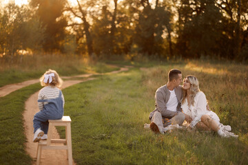Fototapeta premium The parents sit together affectionately, while their toddler, around 2 years old, sits on a stool nearby. Ideal for illustrating love, family values, and serenity in a natural setting.