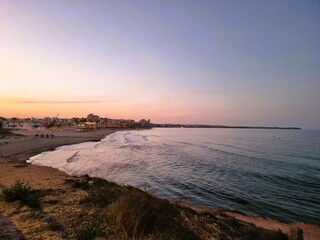 Ein Strand in einer Bucht bei Abendd&auml;mmerung, im Hintergrund leuchten H&auml;user, und ein Landarm ragt ins Meer.