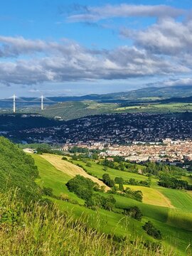 Blick auf das Tal in Frankreich vom Point de Vue du Viaduc de Millau, mit einem Schattenstreifen, der sich durch die Landschaft zieht, und der imposanten Br&uuml;cke im Hintergrund und Wolkenhimmel.