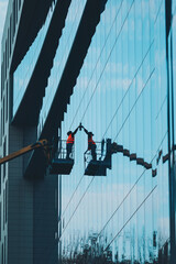 Obraz premium Male window cleaner cleaning glass windows on modern building high in the air on a lift platform. Worker glass high in the air. A worker washes windows in a building from a crane platform