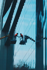 Obraz premium Male window cleaner cleaning glass windows on modern building high in the air on a lift platform. Worker glass high in the air. A worker washes windows in a building from a crane platform