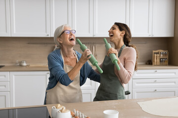 Older woman her young adult daughter hold rolling-pins and singing together in modern kitchen,...