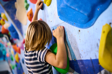 A 6-year-old boy climbing on a vibrant indoor bouldering wall with multicolored holds, engaging in...