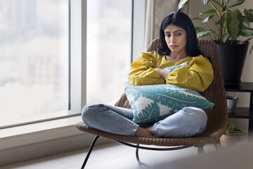 Depressed unhappy young Indian woman sitting in armchair at home, tightening cushion, staring into...