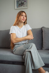 Fototapeta premium Young woman seated on a gray couch showing signs of discomfort while wearing a casual white t-shirt and gray pants in a cozy indoor setting