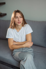 Fototapeta premium Young woman seated on a gray couch showing signs of discomfort while wearing a casual white t-shirt and gray pants in a cozy indoor setting